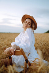 Stylish young girl in a summer white dress and hat posing in a golden wheat field. Fashion,...