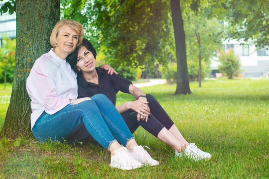 Happy beautiful LGBT couple of mature women in love sitting on the grass in summer park, smiling, looking at camera. Ladies in love spending time together hugging each other. Relationships, friendship