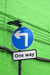 a blue circular road sign with a left turn arrow and notice saying one way against a green brick...