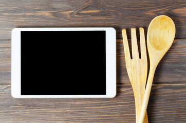 A tablet computer and wooden spoon and fork on a brown background. Copy space. Concept of cooking recipes