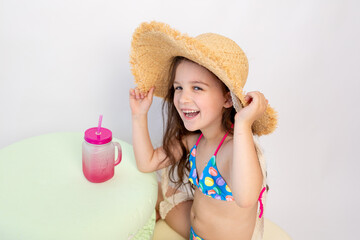 a small smiling girl of 5-6 years old is sitting in a swimsuit and a hat with a cocktail on a white isolated background, a place for text