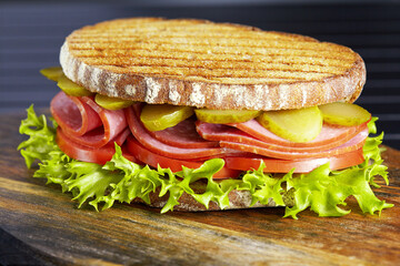 Tasty sandwich with ham, tomatoes, lettuce and sliced pickled cucumbers on a wooden surface. Closeup.
