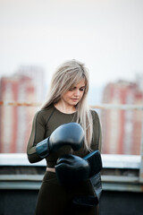 
young adult athletic girl with long blond hair in boxing gloves on the rooftop against the background of the city