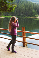 Young Caucasian blond woman making photos on camera near a lake in the mountains.