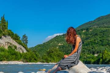 Fototapeta na wymiar An unrecognizable relaxed young Caucasian redhead woman sitting on a boulder with a beer bottle next to the Var river in the French Alps enjoying summer sunshine