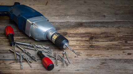 Some handicraft tools like a blue power drill, two red screwdrivers and several silver torx screws are lying on an old and heavy used workbench in a workshop. 