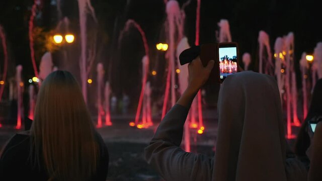 Rear View of people takes photos or filming a video on a smartphone of music fountain with backlight in the evening in a city park