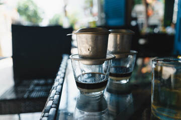 Hot milk coffee dripping in Vietnam style, pouring water over ground coffee contained in Vietnamese Phin Filter on table. Selective Focus.