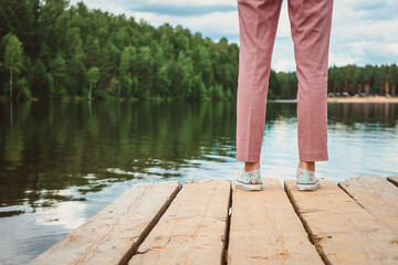 Women's feet stand on the edge of a wooden pier. A girl in sneakers stands with her back to the edge of the docks against the background of the lake and forest