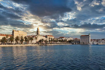 Fototapeta na wymiar Seaside view of the town of Split, Croatia. Riviera with trees and the old town with the saint dominus cathedral belltower peeking above the buildings. Beautiful dramatic sunset lined up with tower