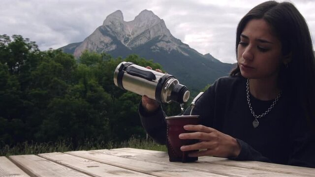 Young woman preparing Argentinean Mate (Mate Argentino) outdoors in a wooden camping table. Girl pouring hot water in mate. Beautiful mountain landscape background
