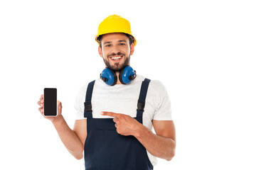 Smiling workman in uniform and protective helmet pointing with finger at smartphone isolated on white