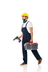Handsome workman in overalls and hardhat holding toolbox and electric screwdriver while walking on white background