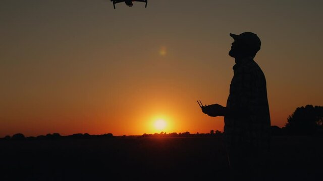 Silhouette of a man at sunset in a field holding a control panel in his hands takes off a drone, looks up