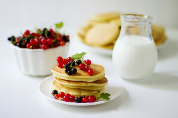 Summer breakfast pancakes with red and black currants in a bowl and milk in a glass jug on a white background.