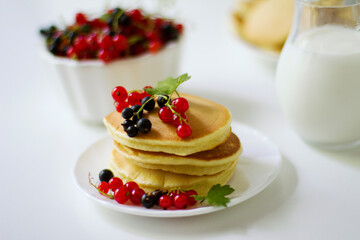 Summer breakfast pancakes with red and black currants in a bowl and milk in a glass jug on a white background.