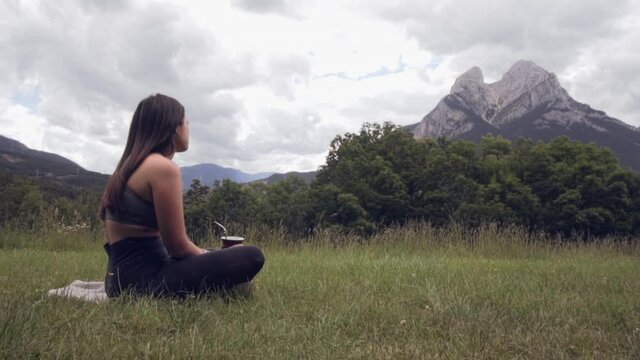 Attractive girl sitting on the grass drinking Argentinean mate (traditional infusion). Relaxed woman taking a break after doing Yoga, admiring the beautiful landscape in Pedraforca, Catalonia
