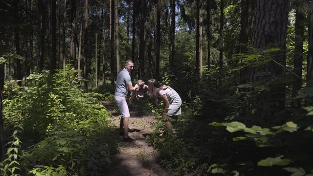 A young father circles the child in his arms in the forest. Next to him, his wife is actively running and enjoying what is happening. Summer day, sun rays. Zoom out of the camera.
