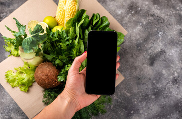 Smartphone in female hand and full box of fresh organic vegetables. Delivery or Order food online concept.