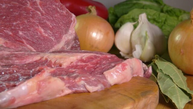 Piece of raw meat is falling on the wooden board on the background of garlic and onions