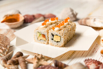 Side view of Asian Sushi in delivery takeaway box on wooden background with Sea shells and stars. Roll with salmon, avocado, sesame seeds, Japanese mayonnaise and red caviar on top.
