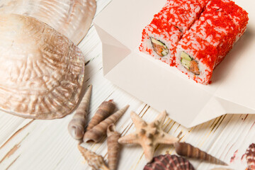 Japanese Sushi in red flying fish roe (Tobiko caviar) on carton delivery takeaway box. California roll with salmon, cucumber, avocado wrapped. Sea shells and stars on wooden background.
