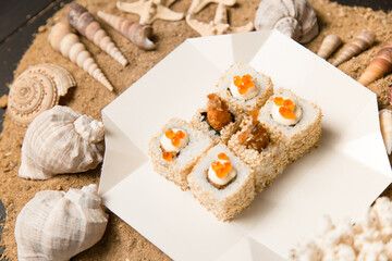 Side view of Asian traditional Ebi Tempura Sushi in delivery takeaway box on sand background with Sea shells. Roll with black tiger shrimp, sesame seeds, Japanese mayonnaise and red caviar on top.
