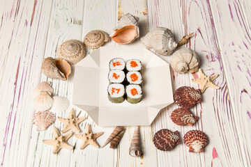 Fototapeta na wymiar Side view of Japanese Sake Maki Roll with salmon and rice wrapped in nori seaweed on white carton delivery box. Sea shells and stars on wooden background. Food delivery creatice concept menu 