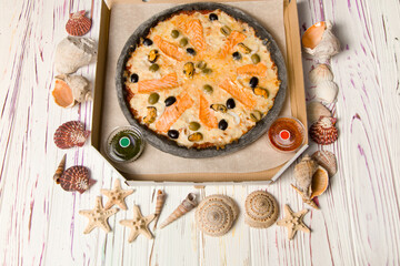 Uncut pizza in delivery box on parchment paper with tomato and basil pesto sauce. wooden background with sea shells and stars. Pizza with cheese, salmon, calamari, mussels and olives on black dough 

