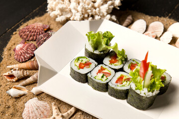 Japanese Futomaki Sushi Roll pieces on carton delivery box. Asian Vegetable Maki roll with tomato, cucumber, avocado, paprika, salad mix on wooden board with ocean coral, sea shells and stars on sand
