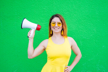 Beautiful happy woman in a yellow dress and sunglasses holds a megaphone. Smiling girl with a loudspeaker in hands on a green background.