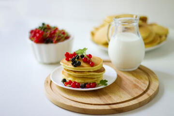 Summer breakfast pancakes with red and black currants in a bowl and milk in a glass jug on a white background.