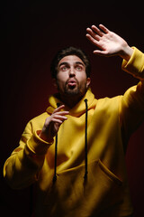 A young man of 25-30 years old in a yellow sweatshirt emotionally pulls his hands up on red wall background. 