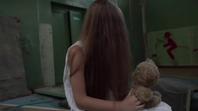 A girl is sitting in a white sundress in an old abandoned school gym. hair on his face, a toy in his hands. Horror.Abandoned house. Mysticism.