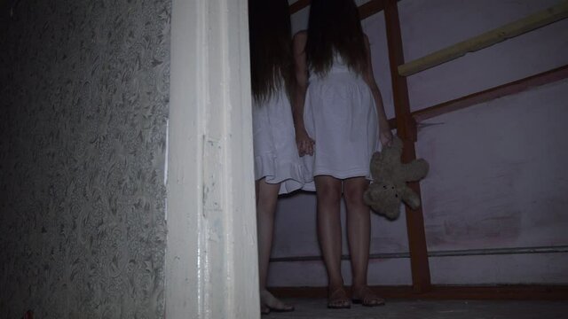 two girls with long black hair and a white dress are standing in an old abandoned room.. The concept of a horror maniac. Halloween. she's holding a Teddy bear,