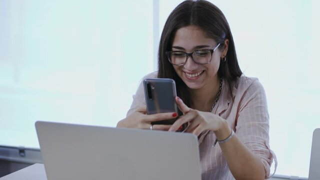 Beautiful young woman typing text message to her boyfriend on smartphone device, checking social media or having video call. Smiling freelancer working in office or from home using laptop pc. 
