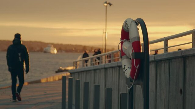 A silhouette man walking past a safety lifebuoy on a modern city harbour, with a blank sea carrying a boat during sunset hour. Focus shift from lifebuoy to man.