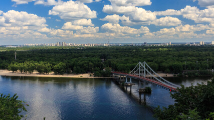 View of the bridge over the river. Sandy beach on the river. Cloudy sky. City summer landscape. Dnipro river in Kyiv on a summer day, Ukraine.  View Of Pedestrian Park Bridge Over The Dnipro River.
