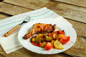 Baked chicken leg with potatoes and tomato on a white plate with a fork on a wooden table.