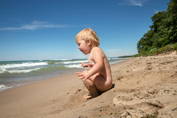 Fototapeta na wymiar Toddler sitting naked on the beach at Indian Dunes National Park enjoying the waves crash in the lake