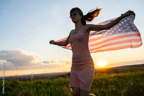 Young woman with USA national flag standing outdoors at sunset. Positive girl celebrating United States independence day. International day of democracy concept.