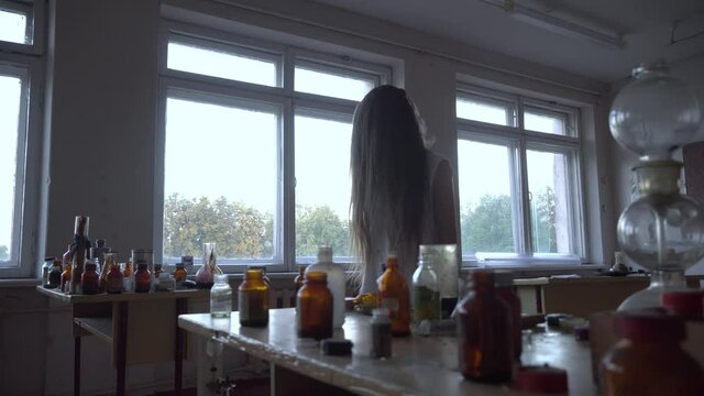 a girl with long black hair and a white dress walks slowly through the dark classroom of the school. The study of chemistry. Against the background of old test tubes and a blackboard.