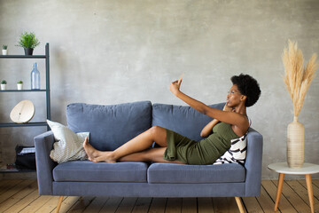 Happy african American young woman sit relax on cozy couch happy to move to new apartment. Smiling black girl rest on comfortable sofa in living room dreaming