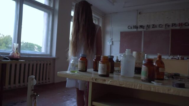 a girl with long black hair and a white dress walks slowly through the dark classroom of the school. The study of chemistry. Against the background of old test tubes and a blackboard.