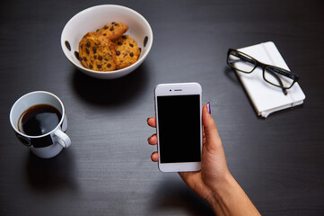 Modern smartphone with blank screen lying flat on coffee table beside some brownies, coffee cup and a notebook