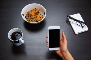 Modern smartphone with blank screen lying flat on coffee table beside some brownies, coffee cup and a notebook