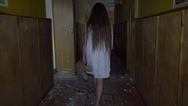 a girl with long black hair and a white dress is walking slowly in a dark corridor. The concept of a horror maniac. Halloween. she is holding a Teddy bear,