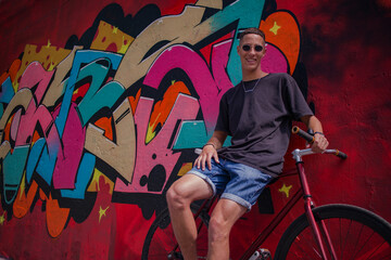Obraz premium a guy with round sunglasses in a dark t-shirt and shorts with a bicycle against a bright wall with graffiti
