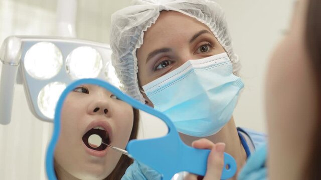 Rear view of Asian girl is lying on dental chair with mirror in her hand and watching female dentist analyzing result of her work using special tool