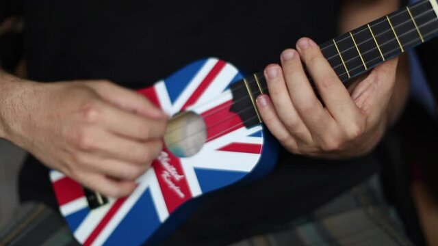 Man Playing Arpeggio And Rhythm On Ukulele Guitar In Close Up And Blurry Background. There Is A British Flag On The Ukule.